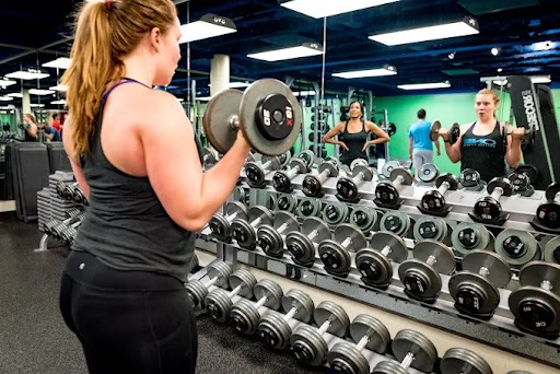 A member curling a dumbbell at the free-weight rack, with other members training reflected in the mirror